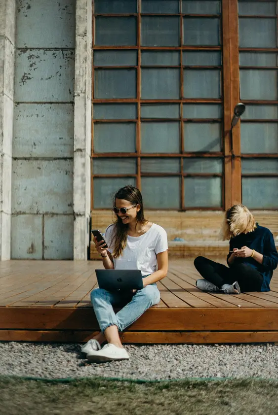 Two people sitting outdoors using laptops and phones, working remotely on a wooden deck.